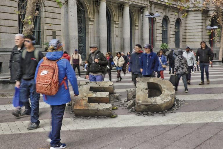 Ilustración: las letras E y D de los fondos de pensiones, de piedra y partidas en pedazos, en medio de una calle peatonal del centro por la que camina gente